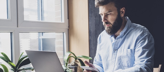 Man working by window in office using wireless digital gadgets for projects. Young professional checking email and sending corporate letters. Office manager typing on laptop and communicating on phone